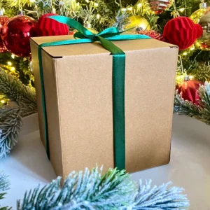 Craft gift box with green ribbon under Christmas tree, surrounded by festive ornaments and frosted branches — Advent candle gift inside for holiday countdown