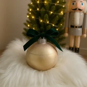 Shiny gold ornament-shaped jar with dark green velvet bow on top, placed on white faux fur with Christmas tree and nutcracker in background