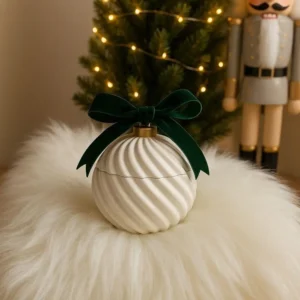 White swirl-patterned Christmas ornament candle jar with green velvet bow, placed on faux fur with festive lights and tree in background