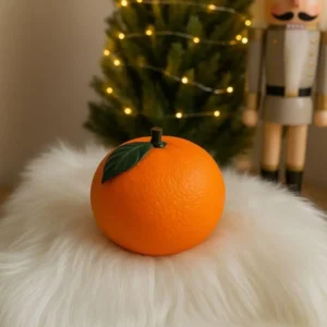 Realistic orange-shaped candle jar with green leaf lid, displayed in cozy Christmas setting with fairy lights and holiday tree