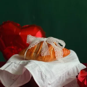 Realistic croissant-shaped aroma candle with a lace bow, placed on white cloth with red heart balloons – a unique and playful Valentine’s Day gift for pastry lovers.