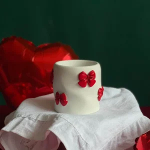 Hand-poured white candle decorated with red bows, perfect romantic gift for Valentine’s Day, surrounded by heart-shaped balloons and festive red-green backdrop.