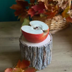 Handmade scented candle shaped like a halved red apple with visible seeds, styled with autumn leaves on a rustic wood stand.