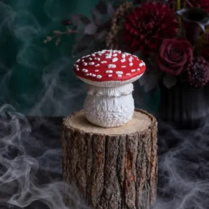 Handmade red and white Amanita muscaria mushroom-shaped scented candle displayed on a rustic wood stand with smoky, moody decor and dark red flowers in the background.