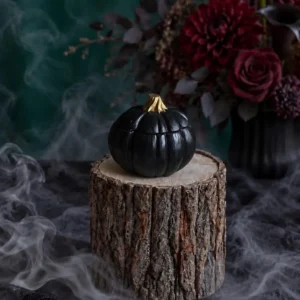 Handmade black pumpkin-shaped scented candle with a gold stem, styled with smoky Halloween decor.