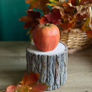 Handmade scented red apple-shaped candle with realistic green and golden tones, styled with autumn leaves on a rustic wood stand.