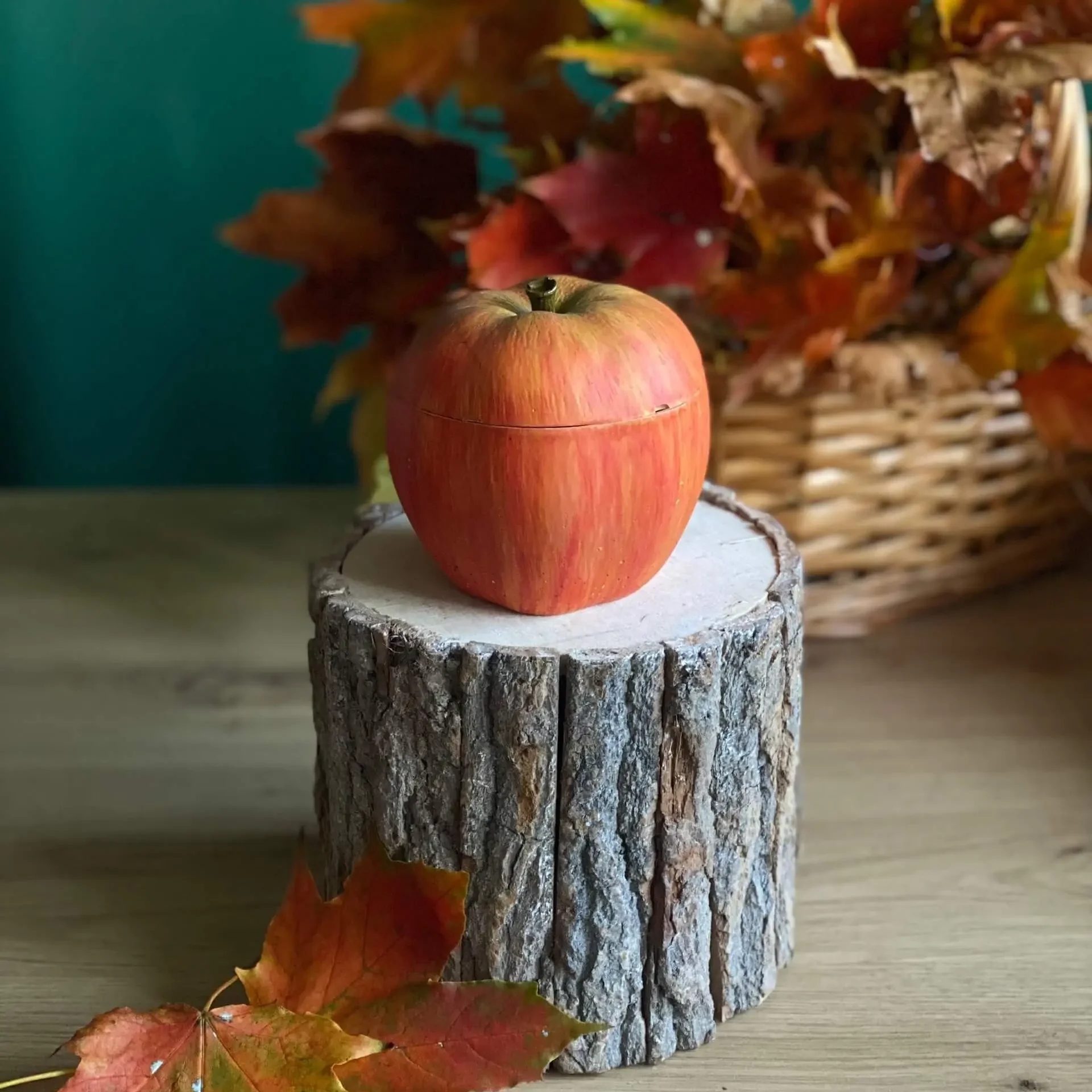 Handmade scented red apple-shaped candle with realistic green and golden tones, styled with autumn leaves on a rustic wood stand.