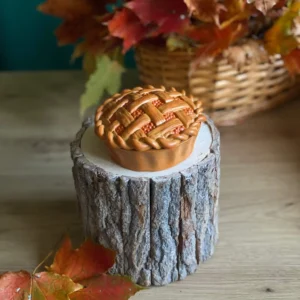Handmade scented pie candle with lattice crust detail, styled on a rustic wood stand with colorful autumn leaves.