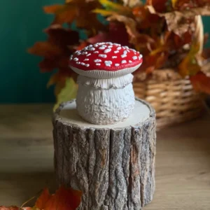 Handmade scented red mushroom candle with white spots, styled on a rustic wood stand with autumn leaves.