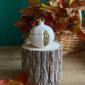 Handmade scented white pomegranate-shaped candle with gold seed details, styled with autumn leaves on a rustic wood stand.