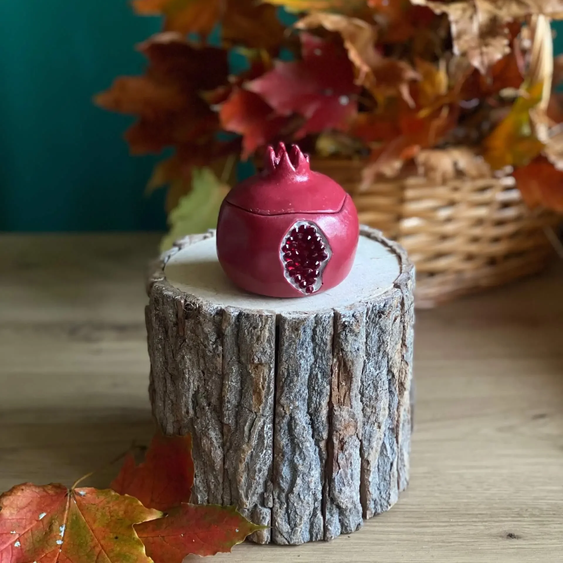 Handmade scented pomegranate-shaped candle in deep red, styled with autumn leaves on a rustic wood stand.