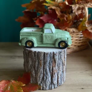 Handmade scented vintage pickup truck candle in soft green, styled on a rustic wood stand with autumn leaves.