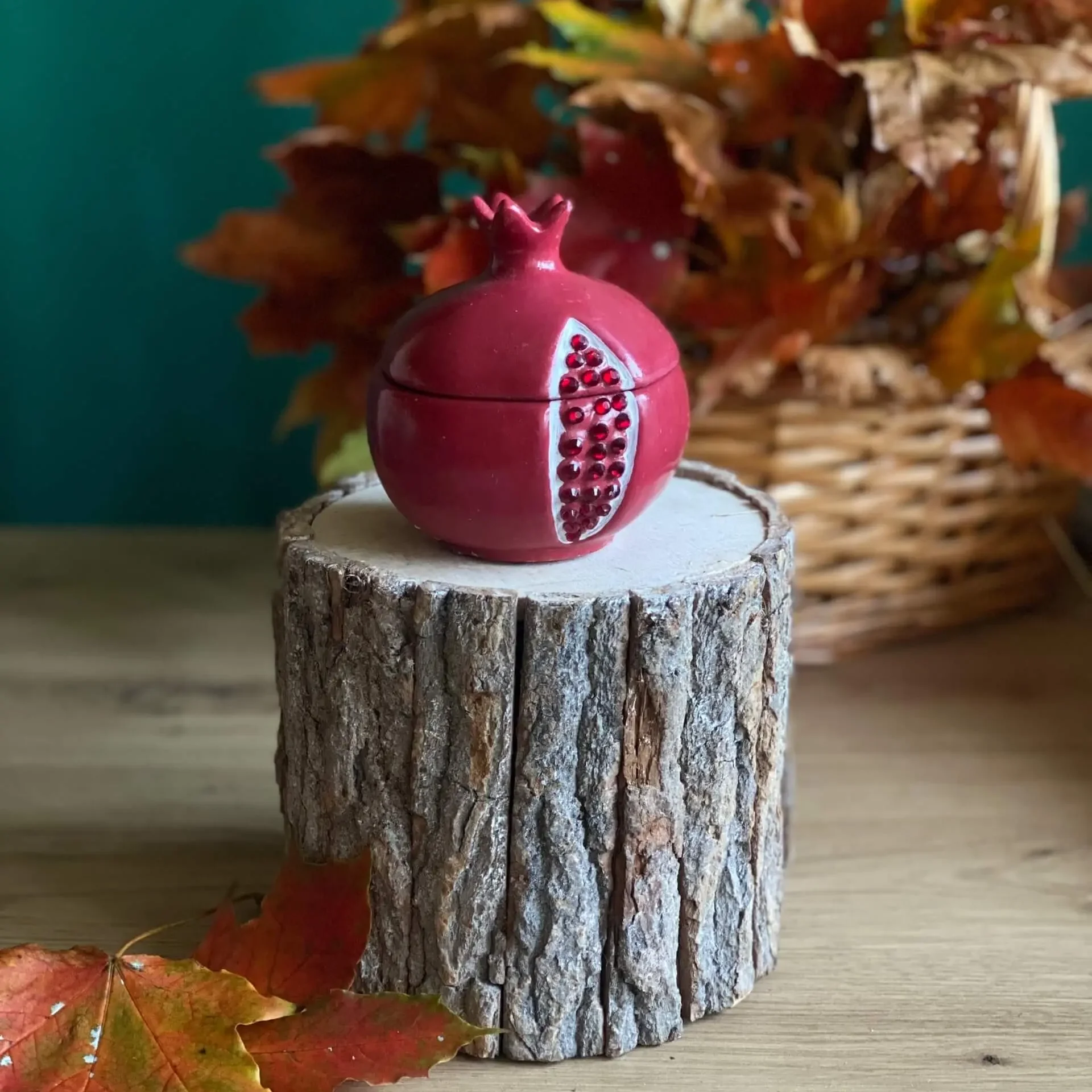 Handmade scented red pomegranate-shaped candle with seed details, styled with autumn leaves on a rustic wood stand.