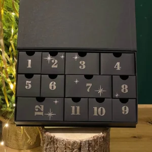 Black 12-day advent calendar with numbered drawers for vintage Christmas ornament-shaped candles, styled by a lit tree.