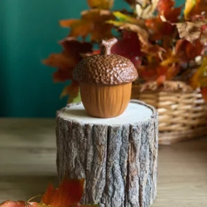 Handmade acorn-shaped scented candle displayed on a rustic wood stand with colorful autumn leaves.