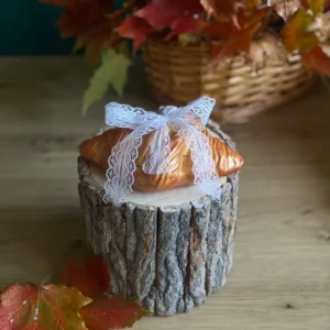 Handmade scented croissant-shaped candle with a white lace bow, styled with autumn leaves on a rustic wood stand.