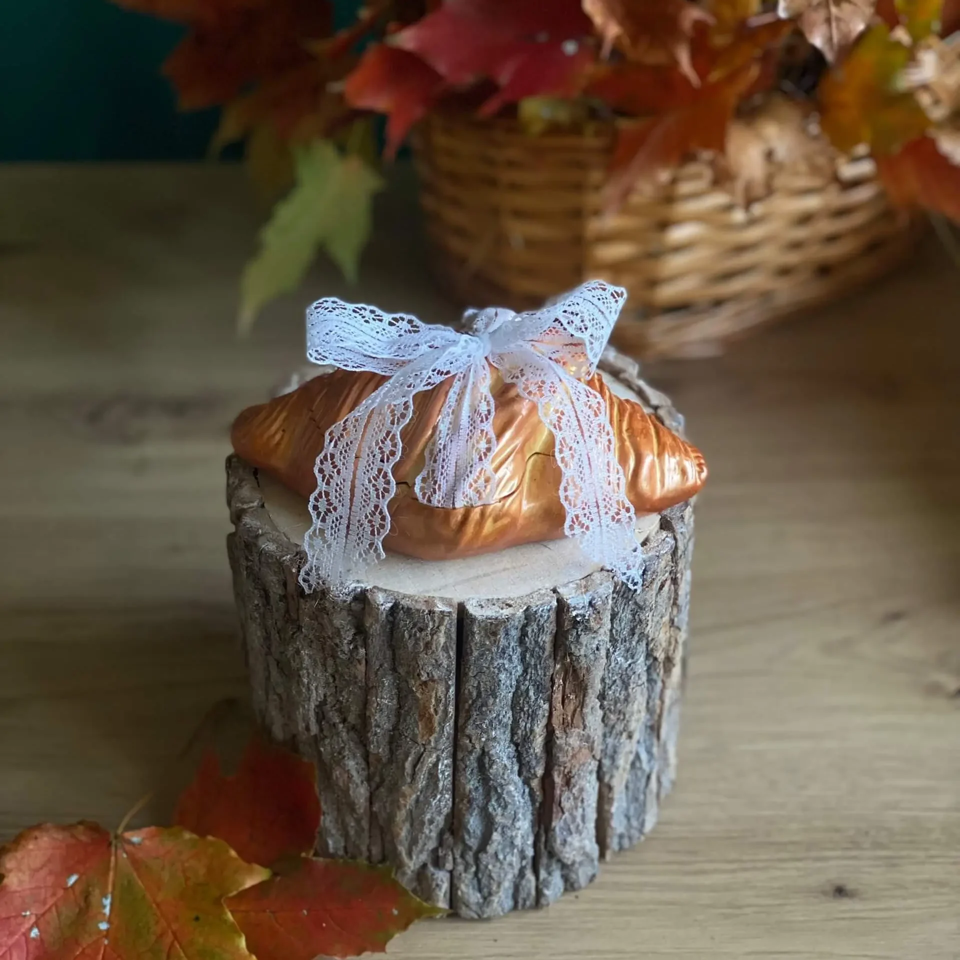 Handmade scented croissant-shaped candle with a white lace bow, styled with autumn leaves on a rustic wood stand.