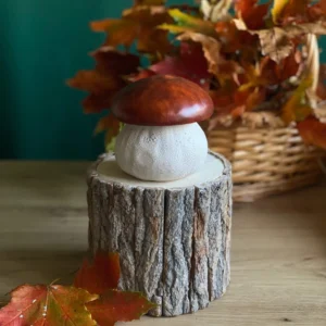 Handmade mushroom-shaped scented candle with a brown cap, styled on a rustic wood stand with colorful autumn leaves.